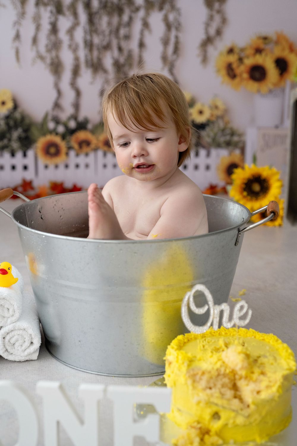 A baby sits in a metal wash tub surrounded by sunflowers checking out her feet during a first birthday cake smash photo session.