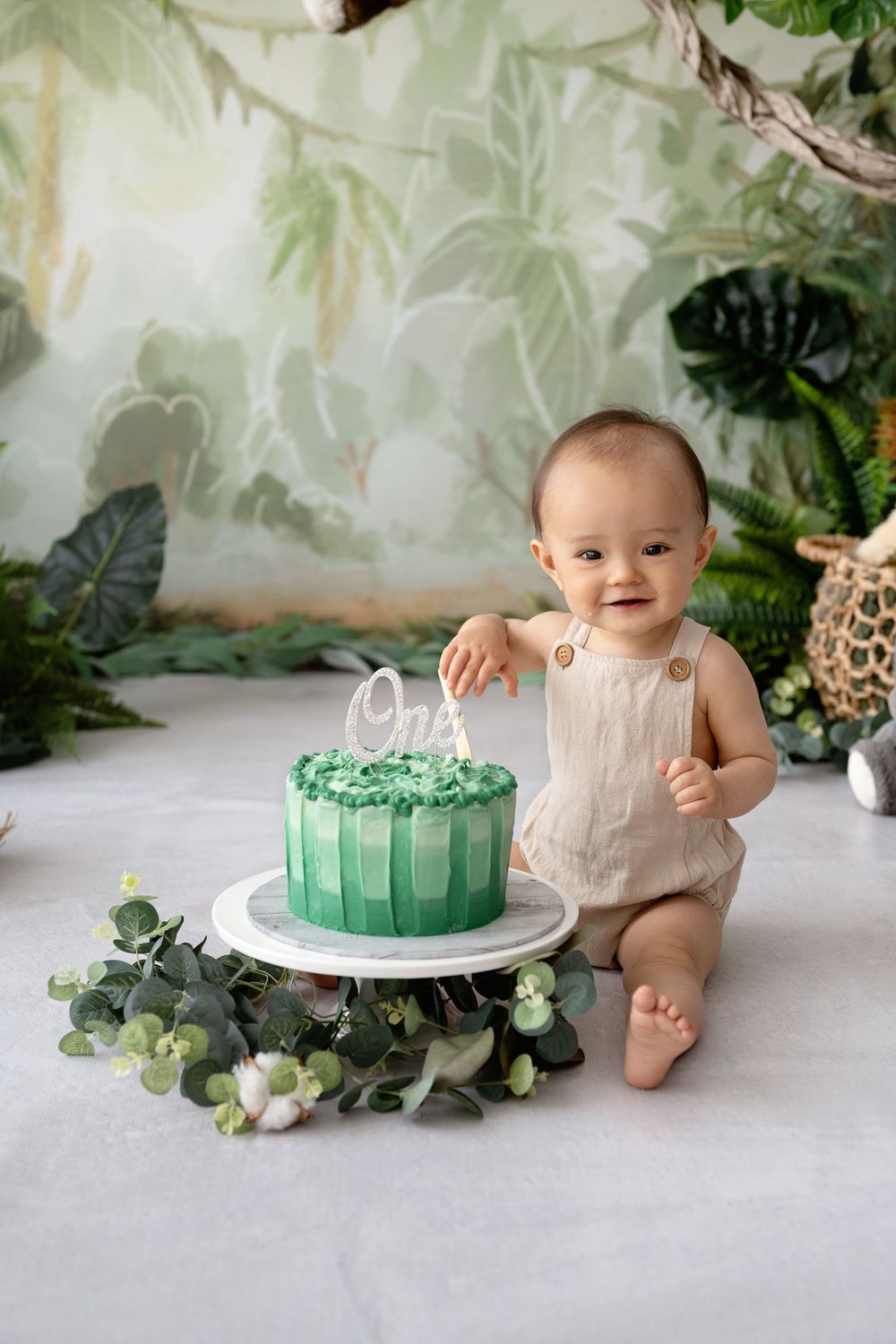 Baby in white outfit sits next to elegant green and white birthday cake decorated with eucalyptus leaves on marble surface.