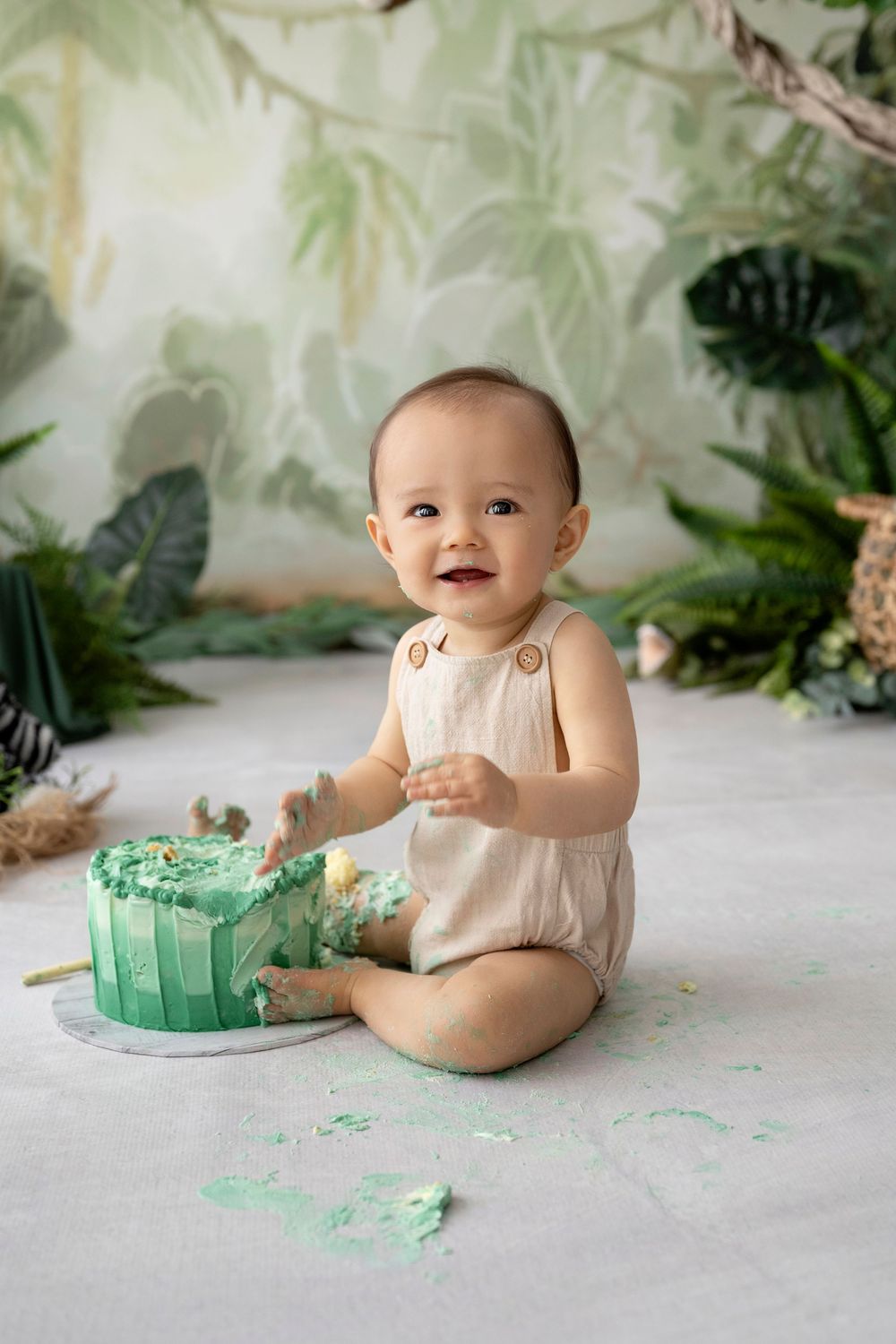 Baby in beige romper sitting with his cake smash decorations and frosting during milestone photo session.