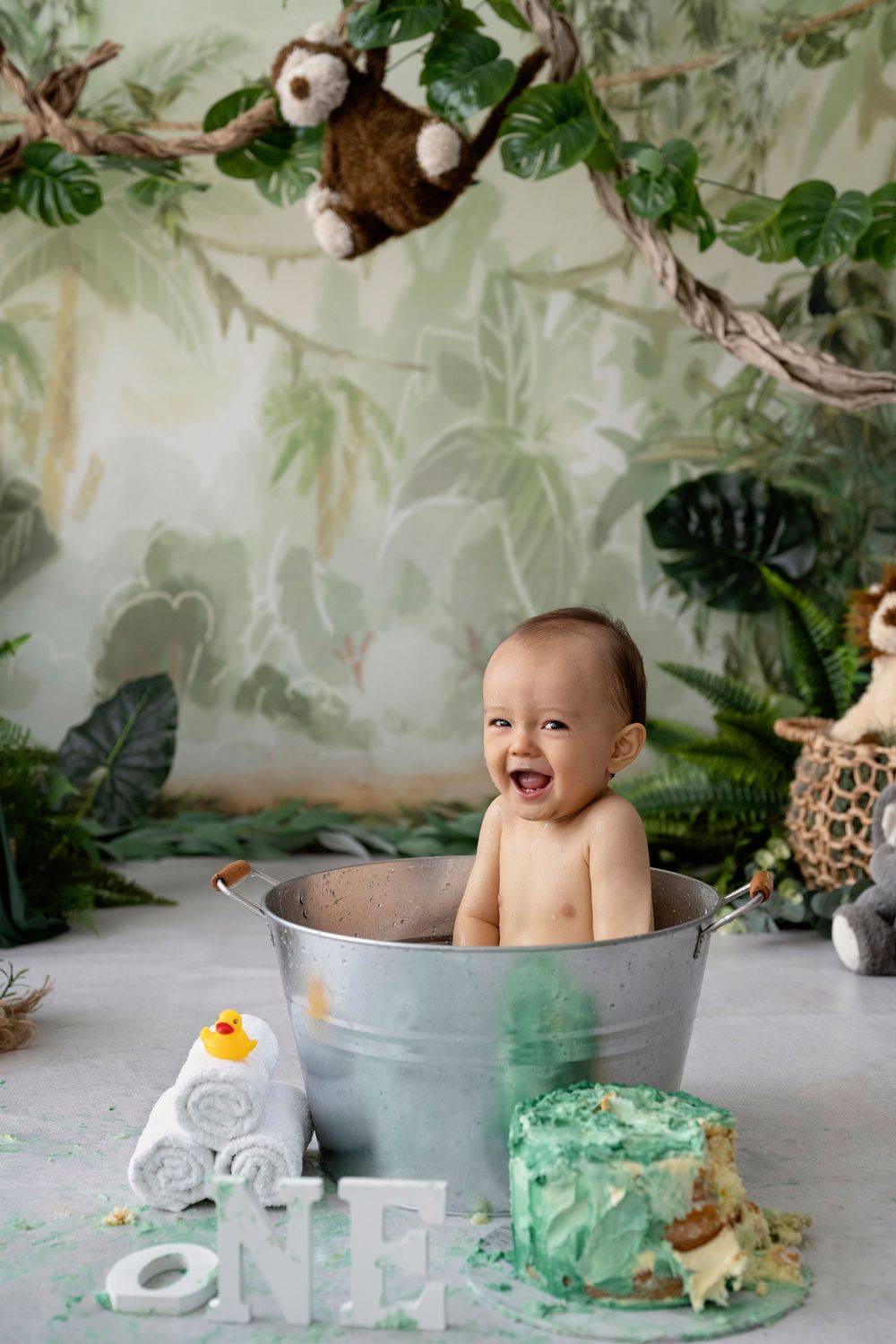 A baby sits in a metal washtub for a cake smash photo session with jungle-themed decorations and props.