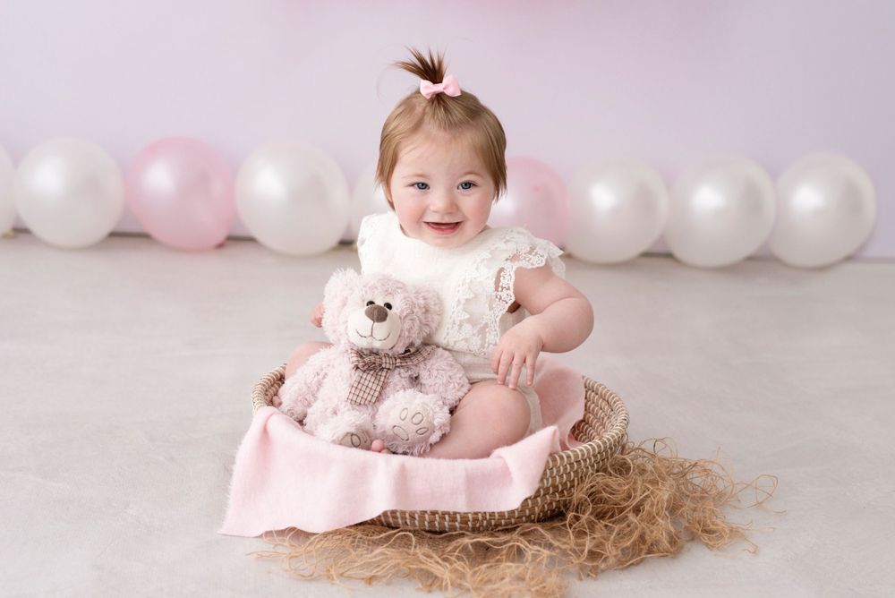 Baby in white dress sits on pink blanket with teddy bear, surrounded by pink and white balloons.