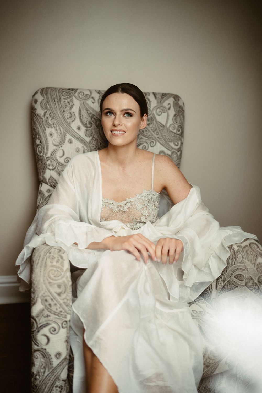 Elegant bride wearing a delicate lace negligee and sheer robe relaxes in an ornate gray armchair.