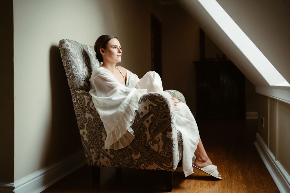 Bride in white dress sits contemplatively in vintage chair by window light.