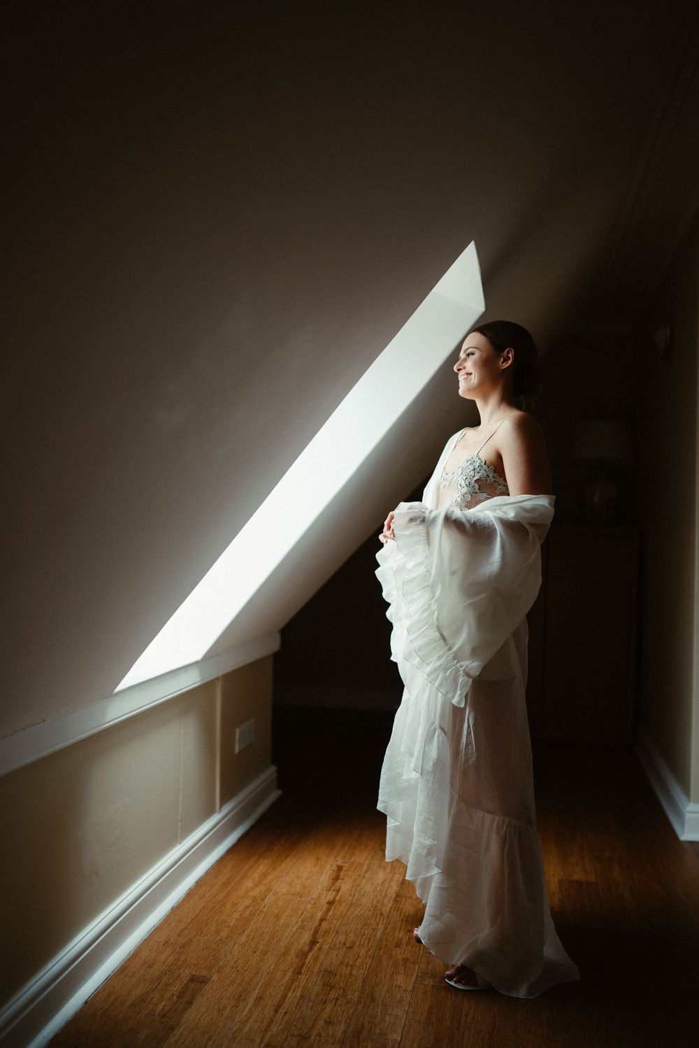A bride in a white gown stands by a slanted window in an attic room with dramatic natural lighting.