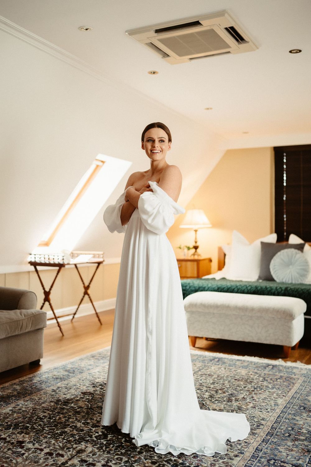 Bride in flowing white off-shoulder gown poses in sunlit room with modern decor.