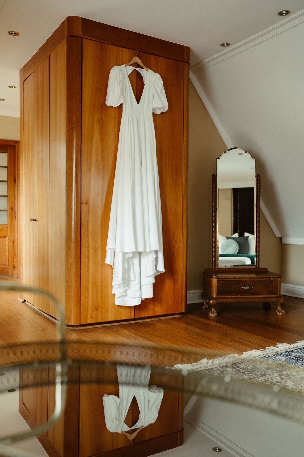 Elegant white wedding dress with puff sleeves hanging on wooden wardrobe with reflection on floor.