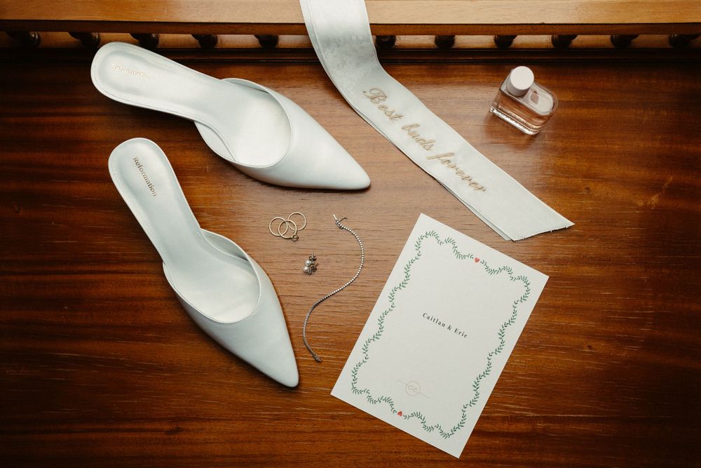 Wedding accessories including white heels, invitation and jewelry arranged on wooden surface.