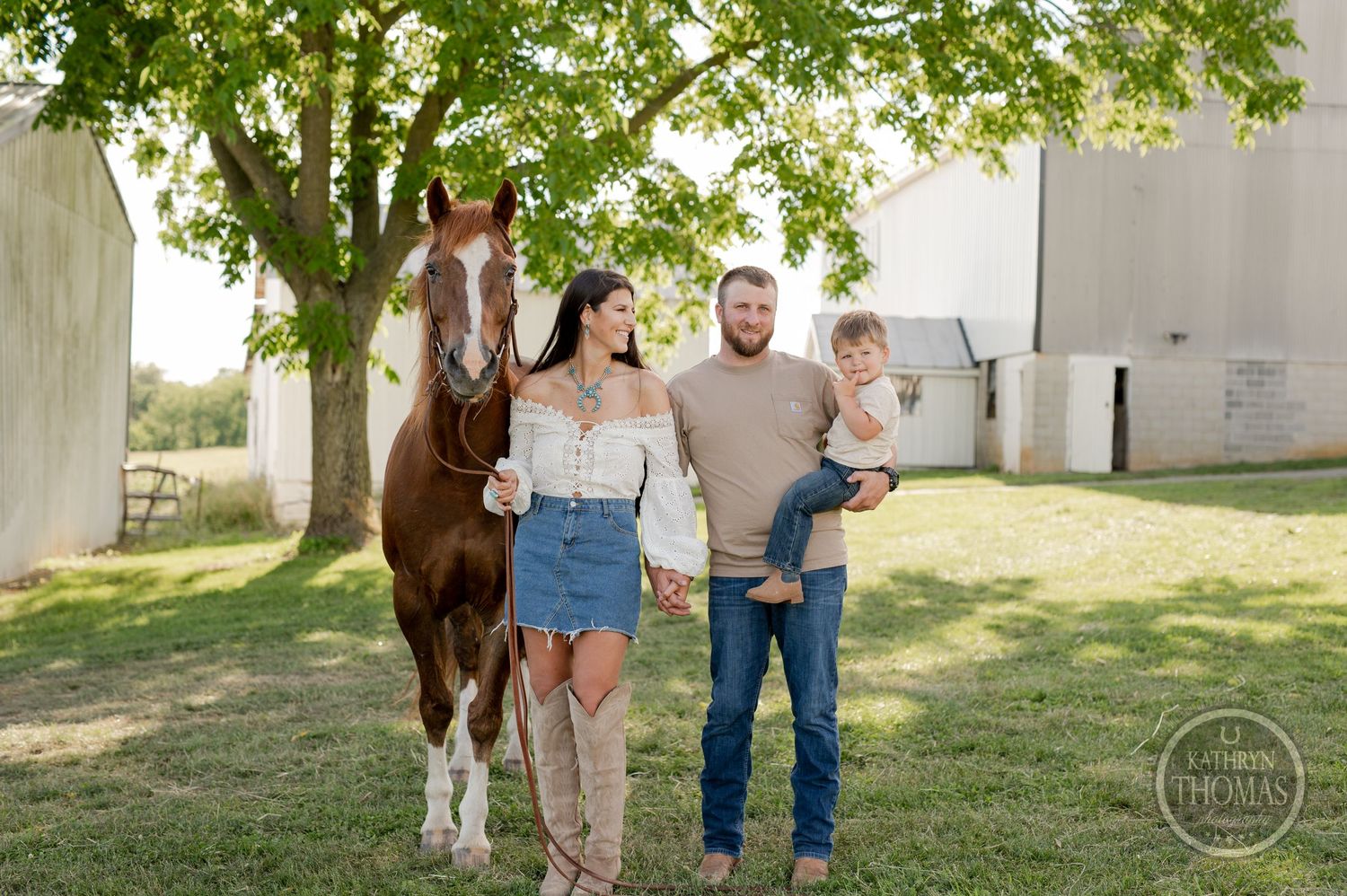 Family poses together with brown horse in sunny outdoor setting near white barn.