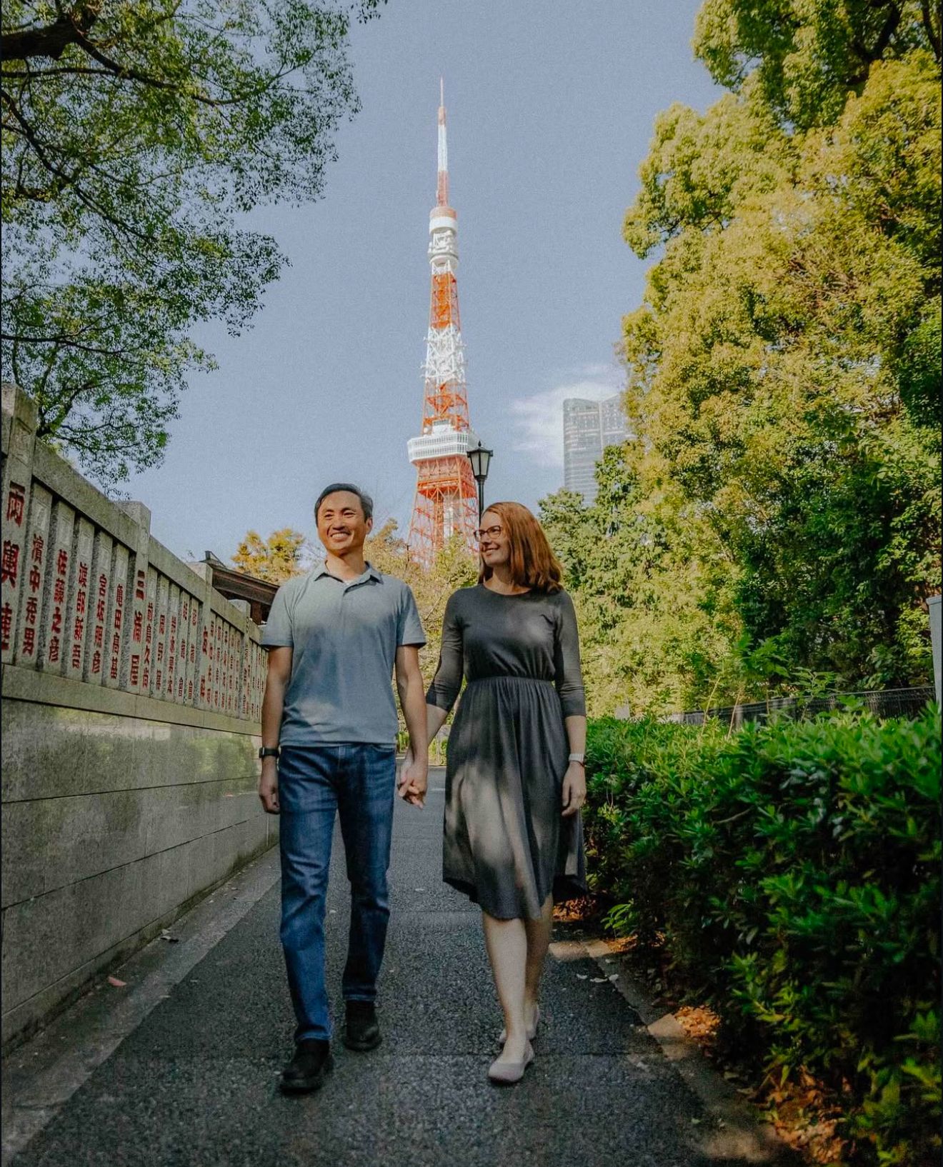Two people walking together on a path with Tokyo Tower and lush green trees in the background on a sunny day.