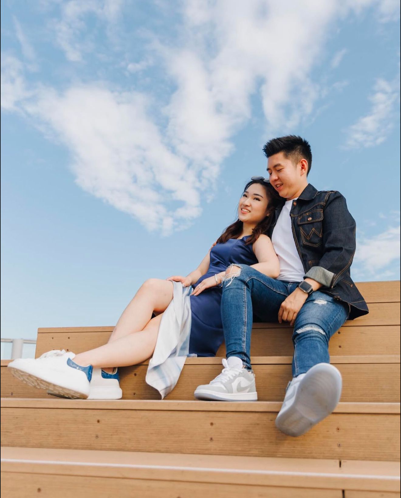 A couple sits casually on wooden steps under a blue cloudy sky, wearing jeans and white sneakers.