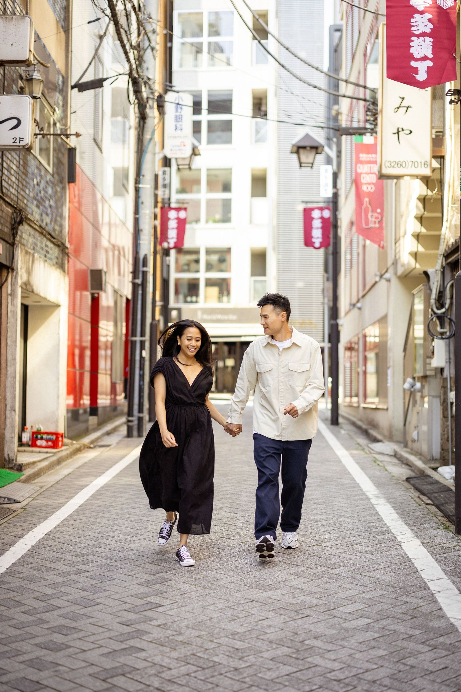 A couple walks hand in hand down a narrow urban alley with Japanese signage and buildings in Tokyo.