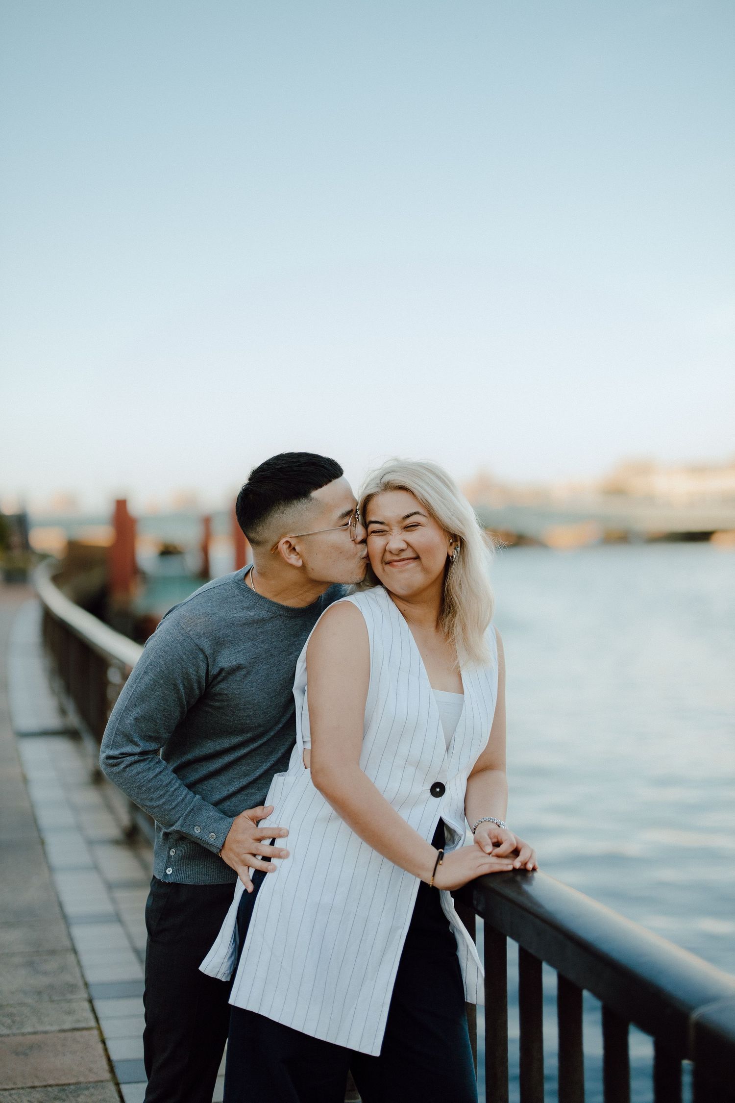 Romantic couple shares tender moment along waterfront railing at sunset with city skyline in background.