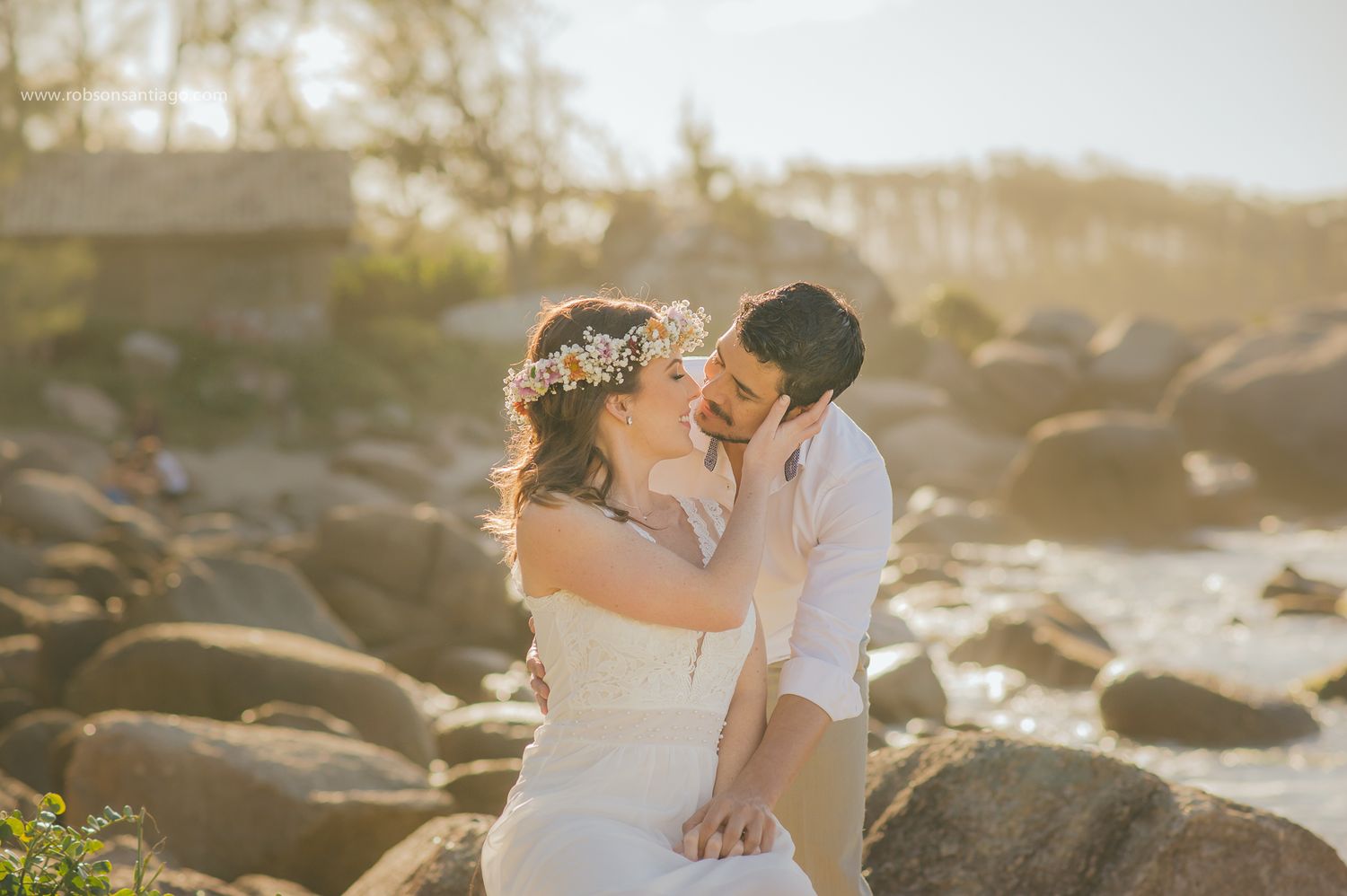 A romantic wedding couple embraces on a rocky beach at sunset with golden light glowing in the background.