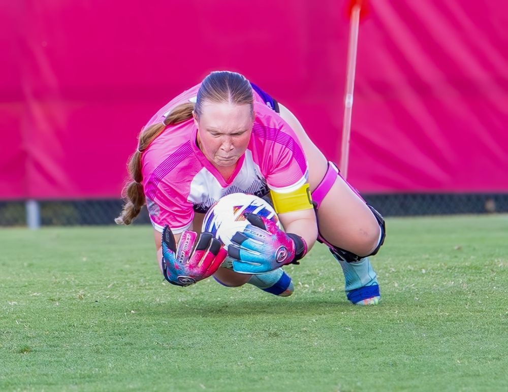 Soccer goalkeeper in pink jersey makes diving save during match on grass field with magenta background.