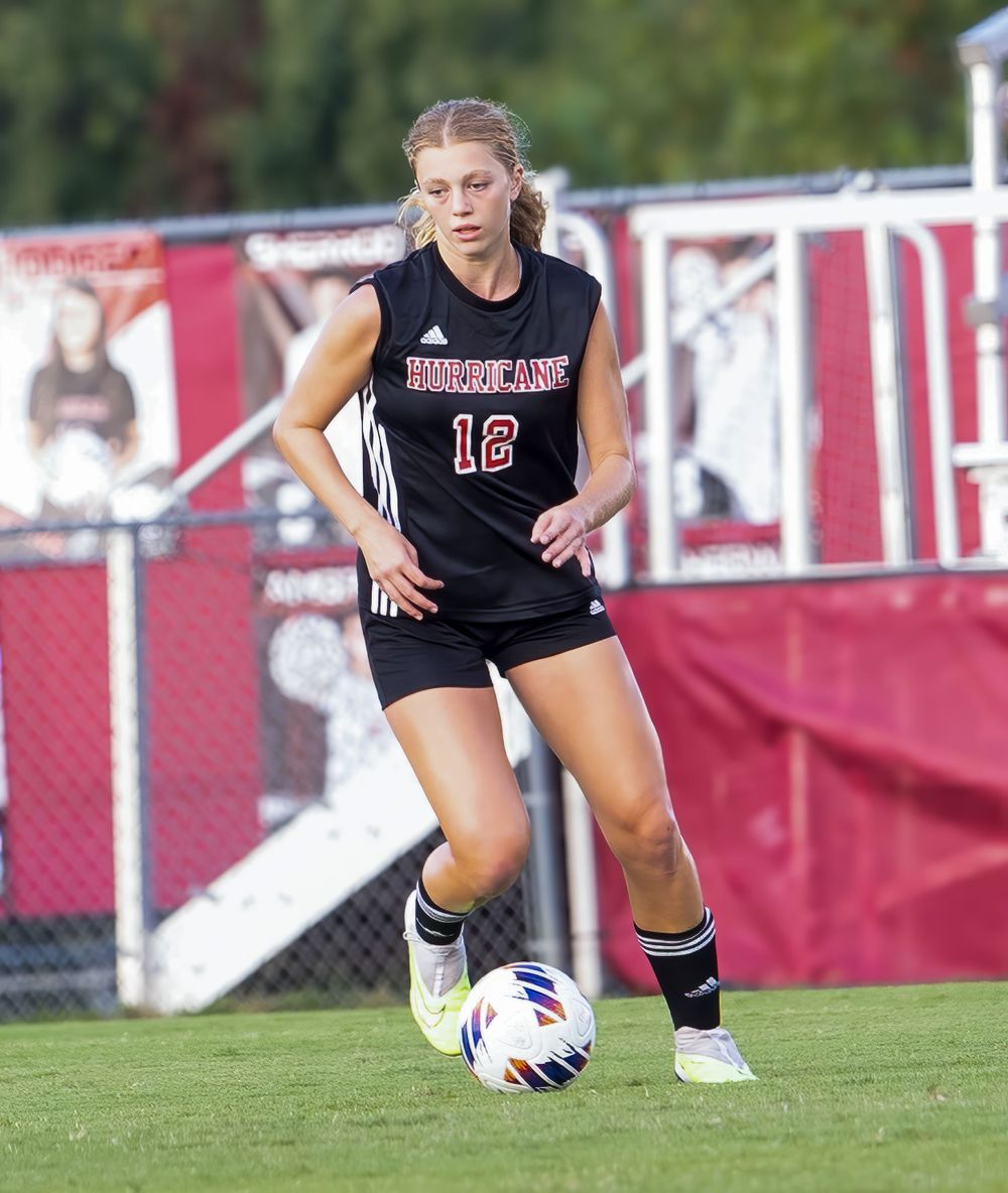 Soccer player in black uniform dribbling ball on field with red fence in background.