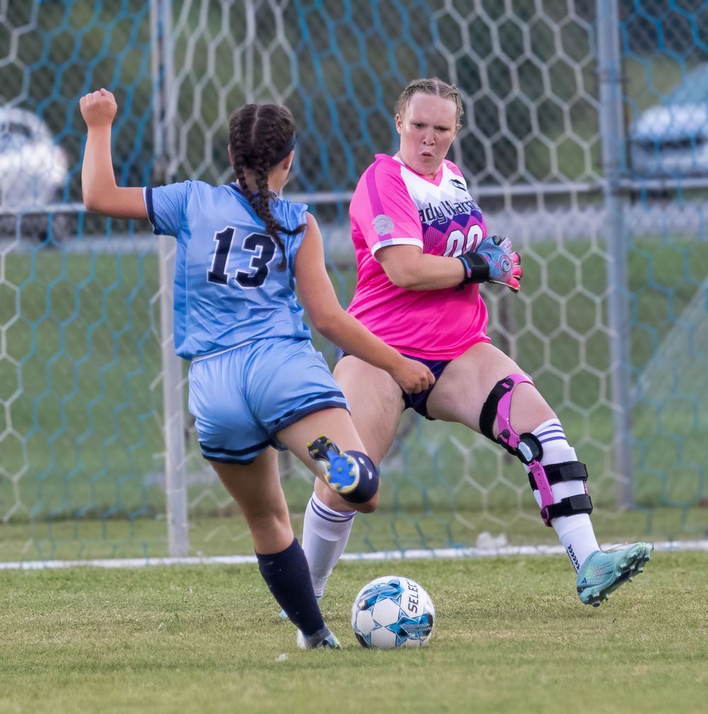Players battle for ball during soccer match with goalkeeper in pink jersey defending goal.