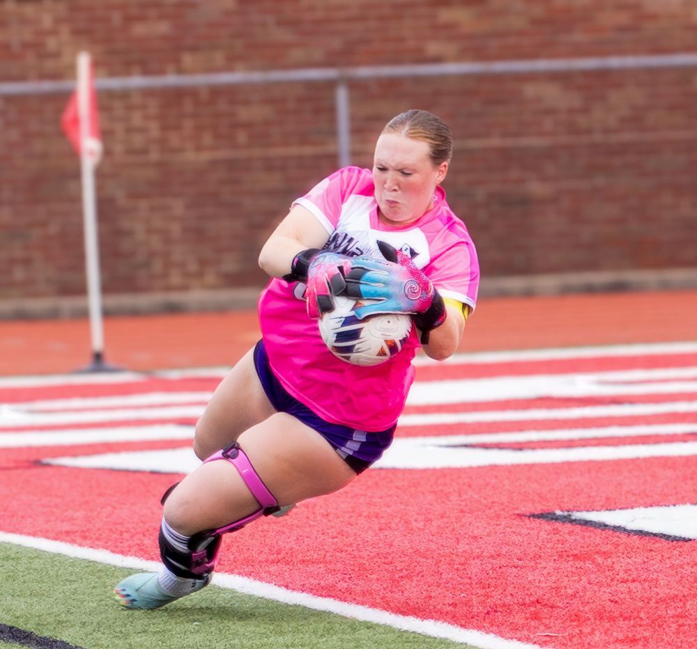 Goalkeeper in pink jersey makes diving save during soccer match on red turf field.