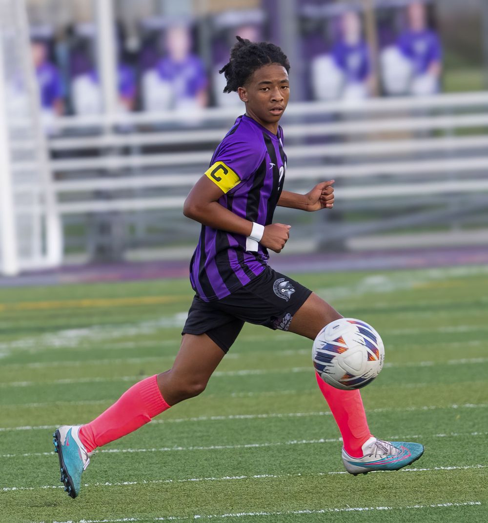 Soccer player dribbles the ball while wearing purple and black striped uniform with red socks.