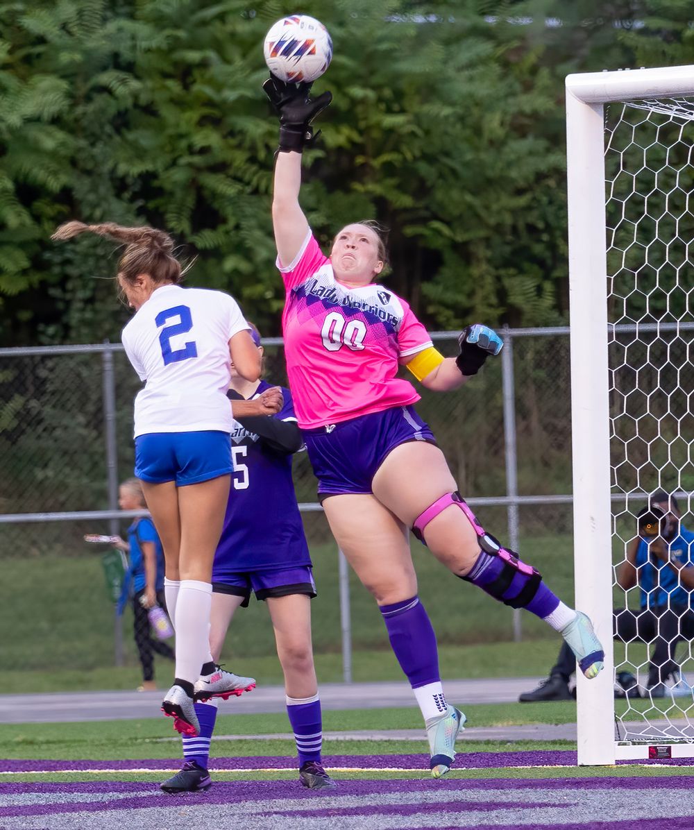 Goalkeeper in pink jersey jumps to make a dramatic save at the goal post during a soccer match.