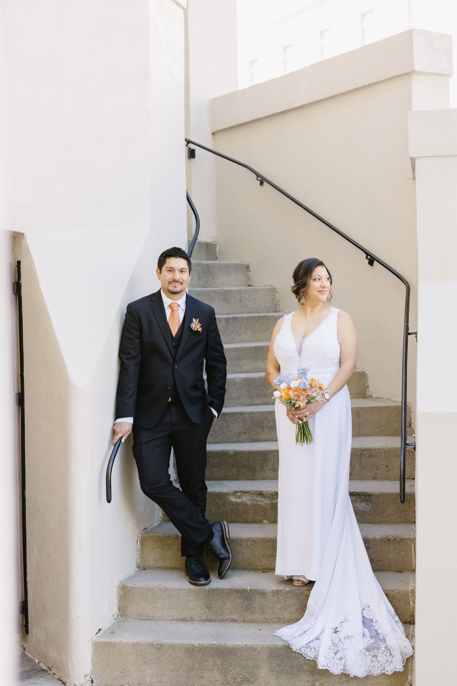 A couple sits on concrete steps against a white wall, with the bride holding a colorful bouquet.