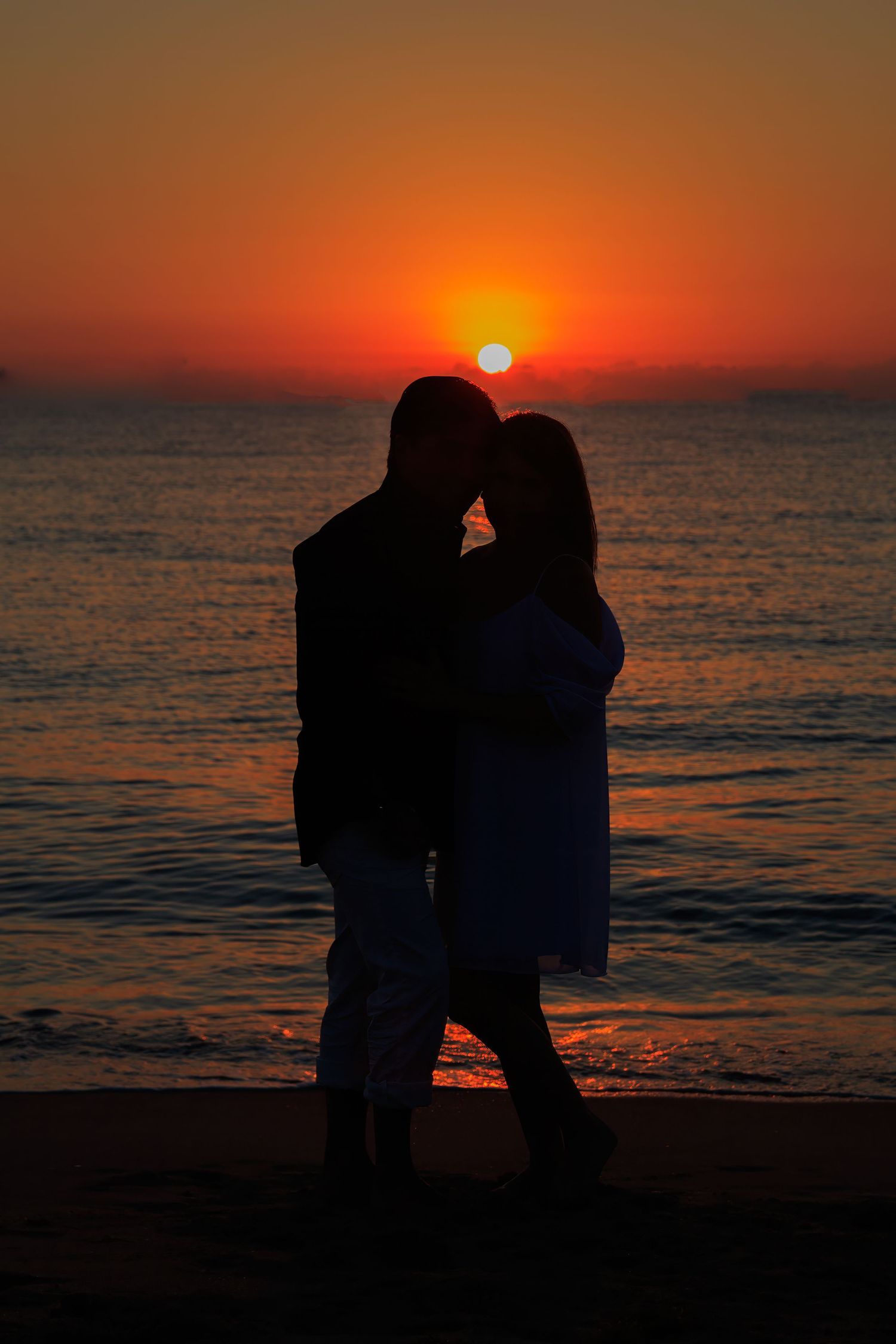 Silhouetted couple embracing on beach during dramatic orange sunset over ocean.