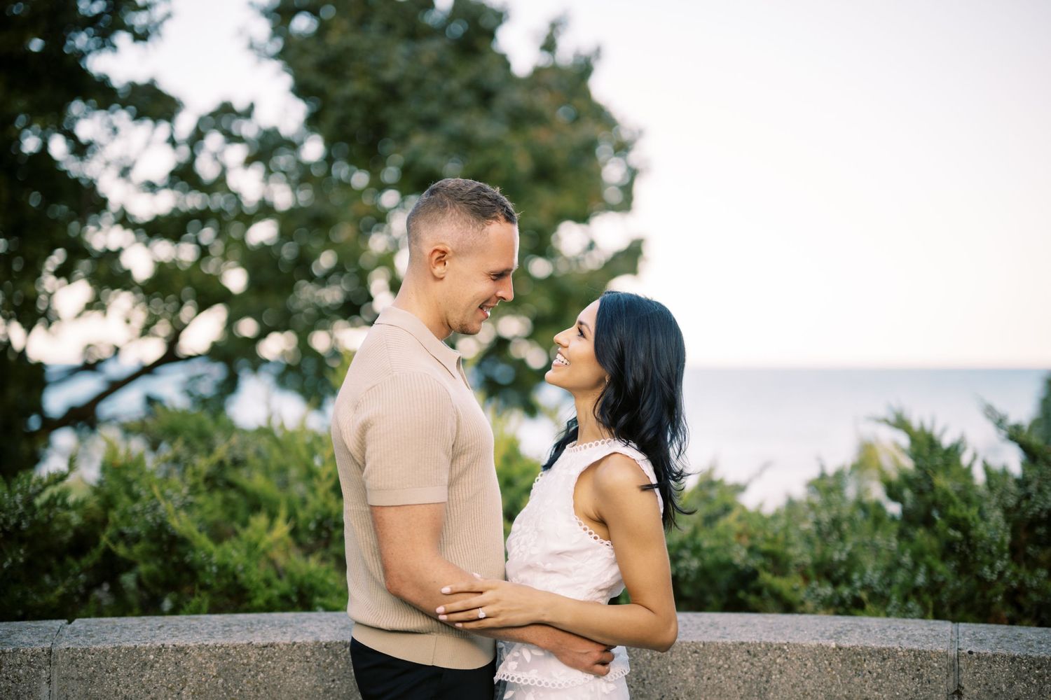 A romantic couple embraces while standing on a coastal overlook at sunset with trees and ocean in the background.