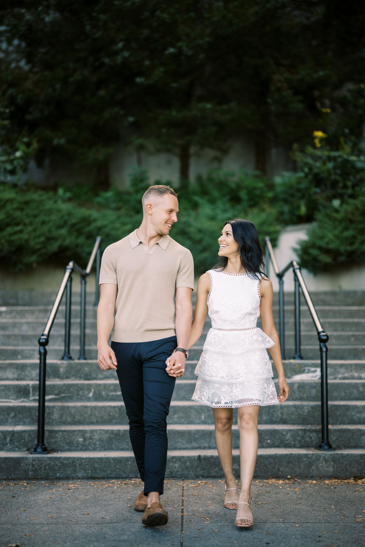 A couple holding hands while walking down outdoor stone steps surrounded by greenery in casual dressy attire.