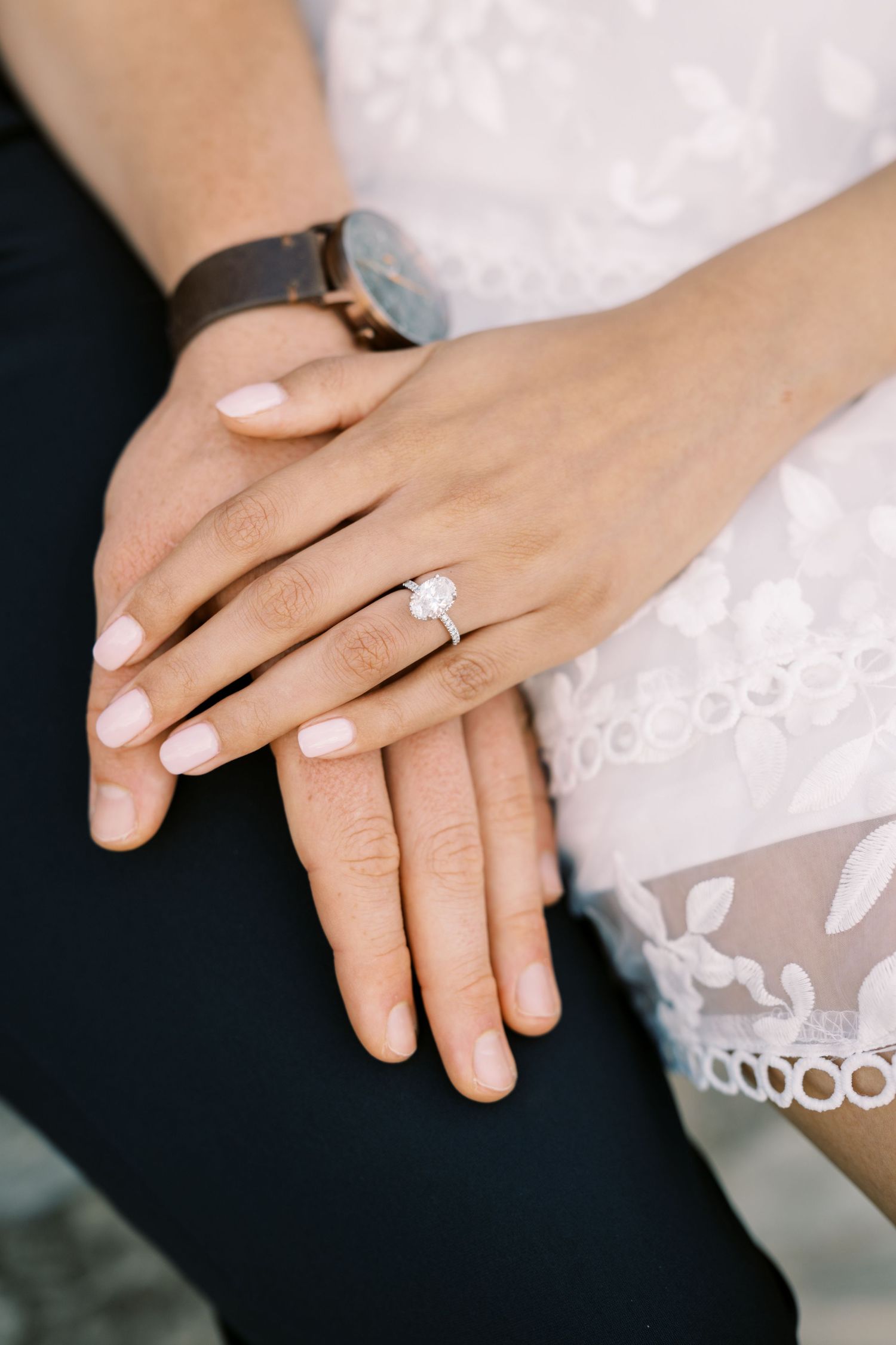 Close-up of hands showing elegant engagement ring with white manicured nails and lace dress details.