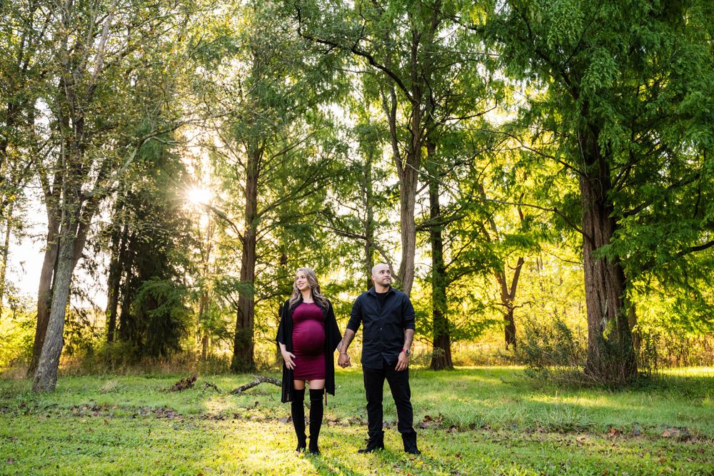 A couple walks together through a sunlit forest path with tall trees and green grass.