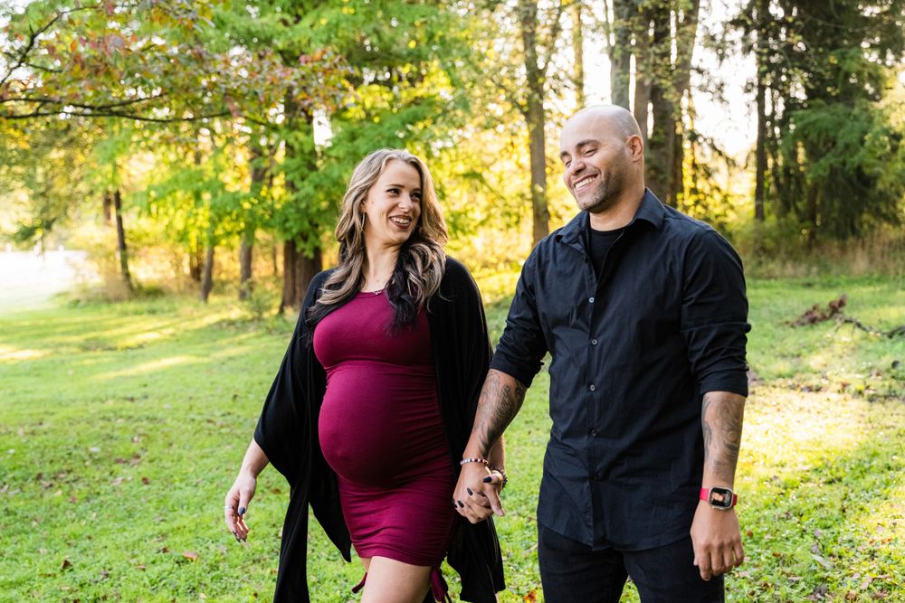 A couple walks together in a sunlit wooded area during an autumn maternity photo session.