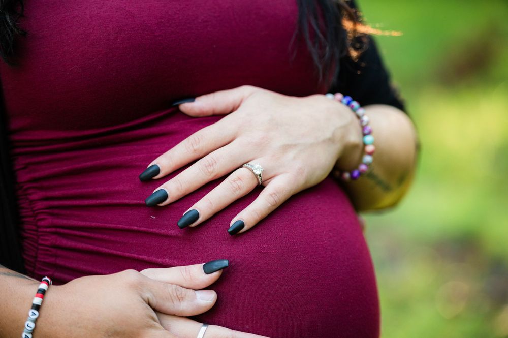 Close up of hands resting on a pregnant belly wearing a burgundy maternity dress with dark blue painted nails.
