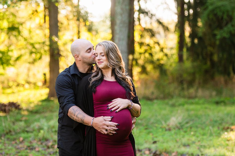 A couple in a forest setting during golden hour with a burgundy dress and black shirt embracing in warm sunlight.