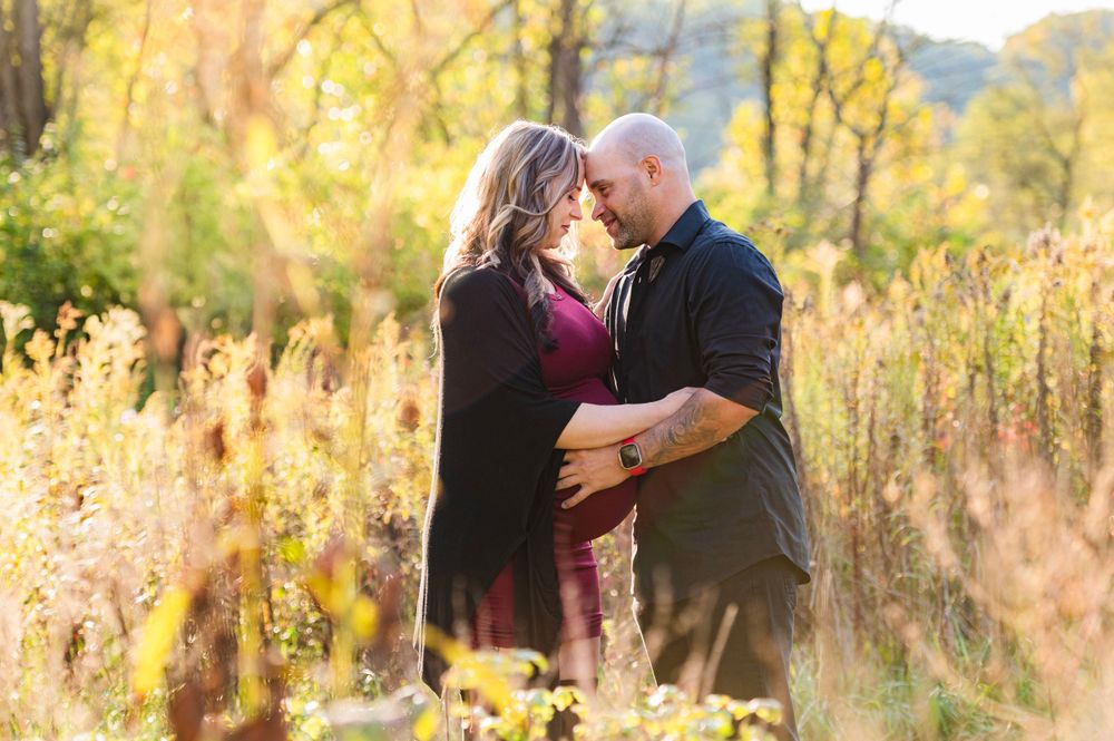 A couple embraces in a sunlit autumn field with tall golden grass and trees glowing in the late afternoon light.