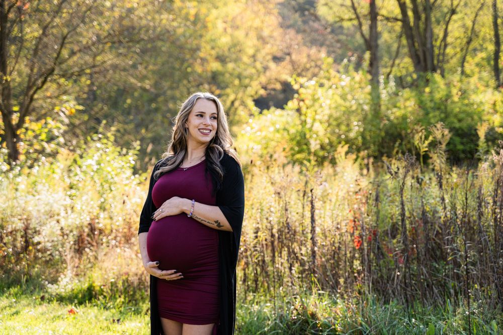 A maternity photo shows an expectant mother in a burgundy dress standing in a sunlit meadow during golden hour.