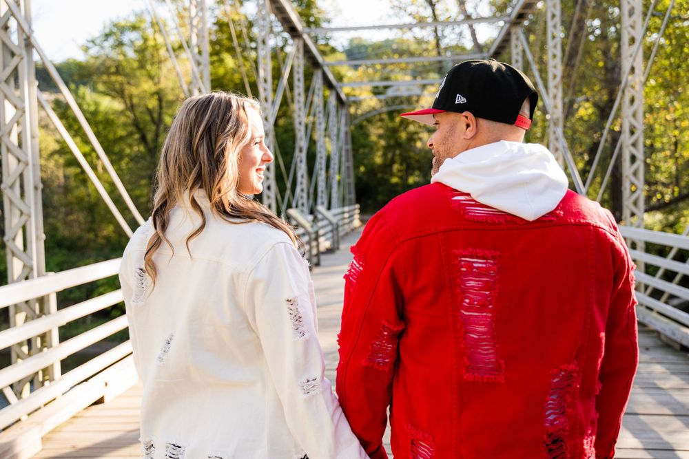 Two people in white and red hoodies walk together on a rustic suspension bridge surrounded by autumn trees.
