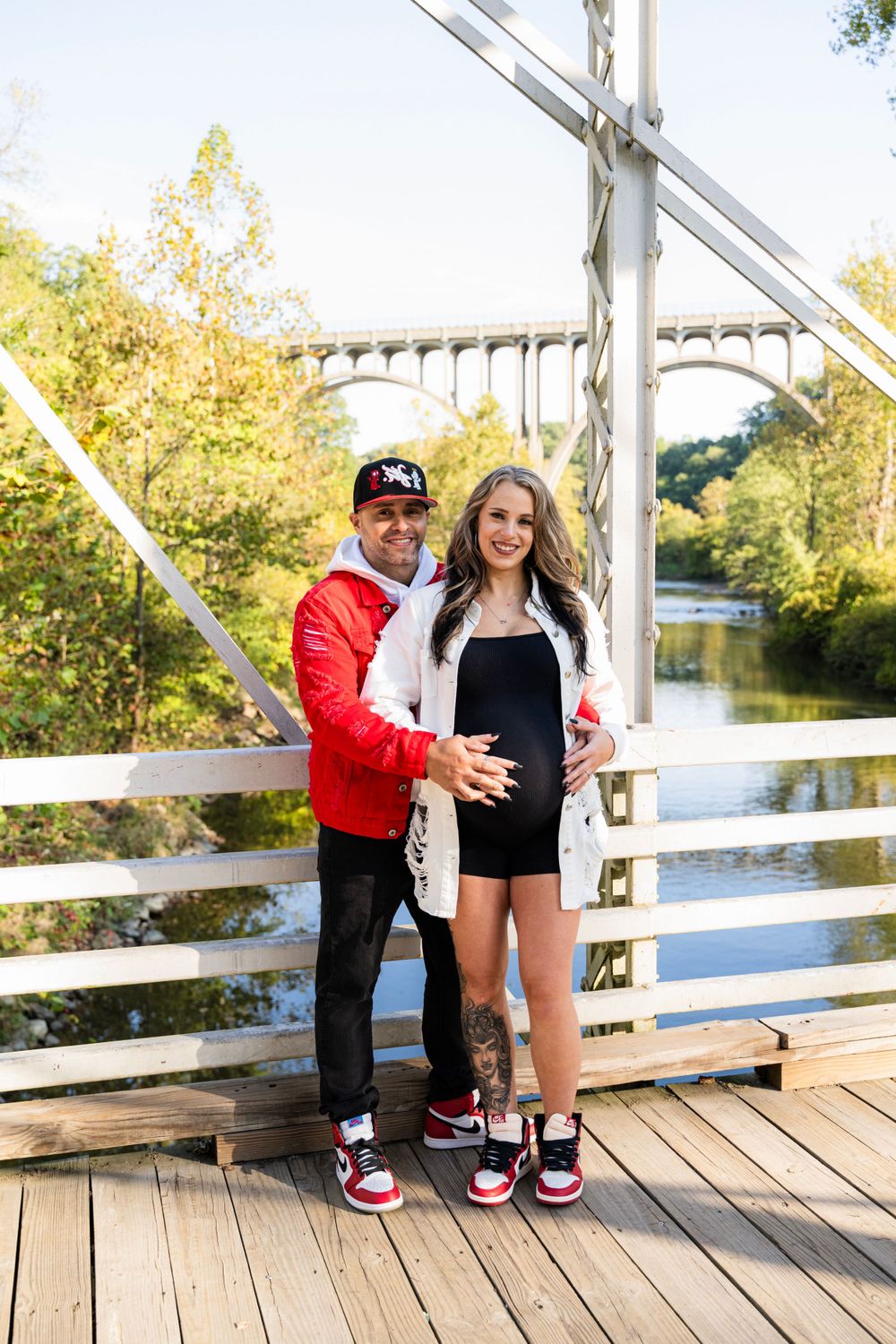 Couple in matching red and white outfits pose together on a wooden bridge with an arched bridge visible in background.