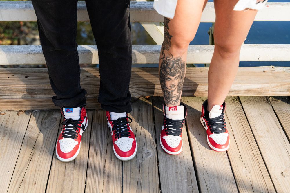 Two pairs of matching red and white sneakers worn by a couple standing on wooden deck, showing tattoo on leg.