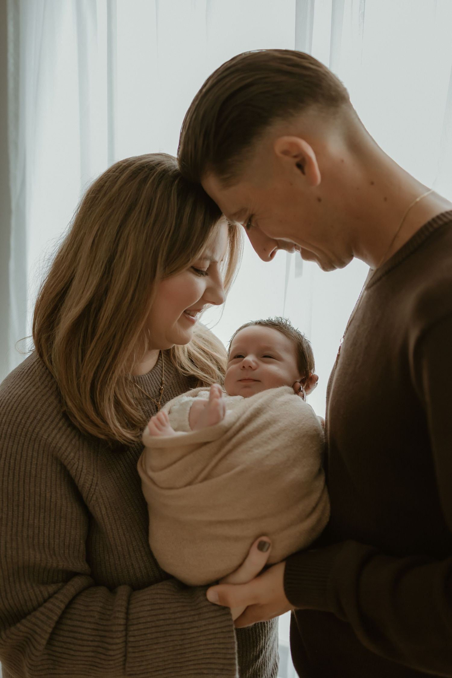 A couple shares a tender moment with their newborn baby by a window with soft white curtains in the background.