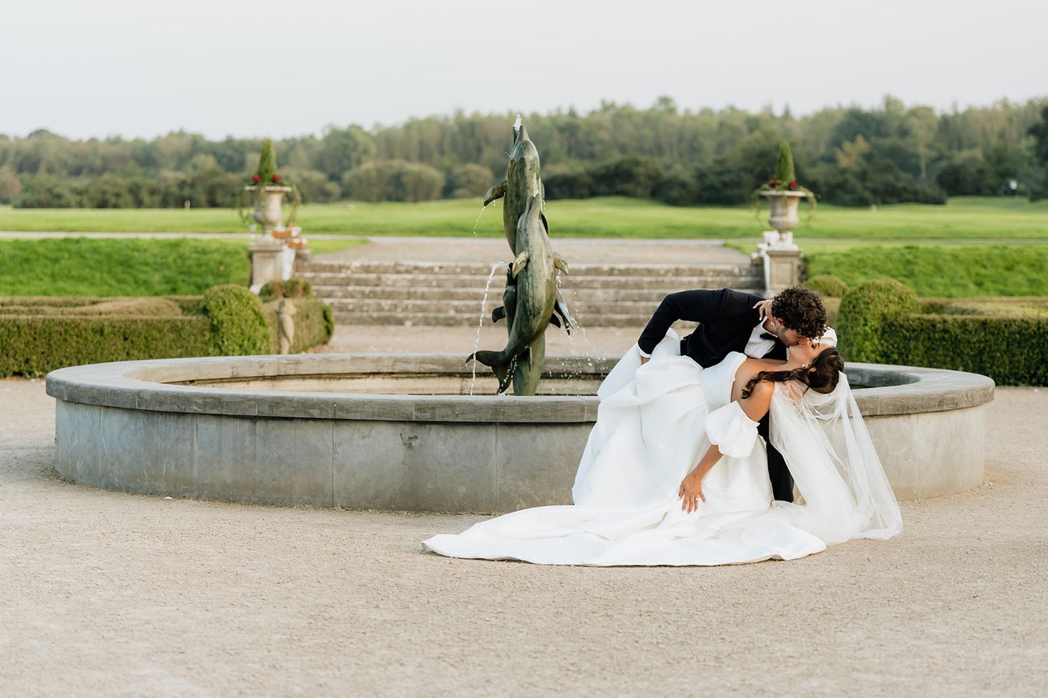 Romantic dip pose by ornate fountain in formal garden setting.