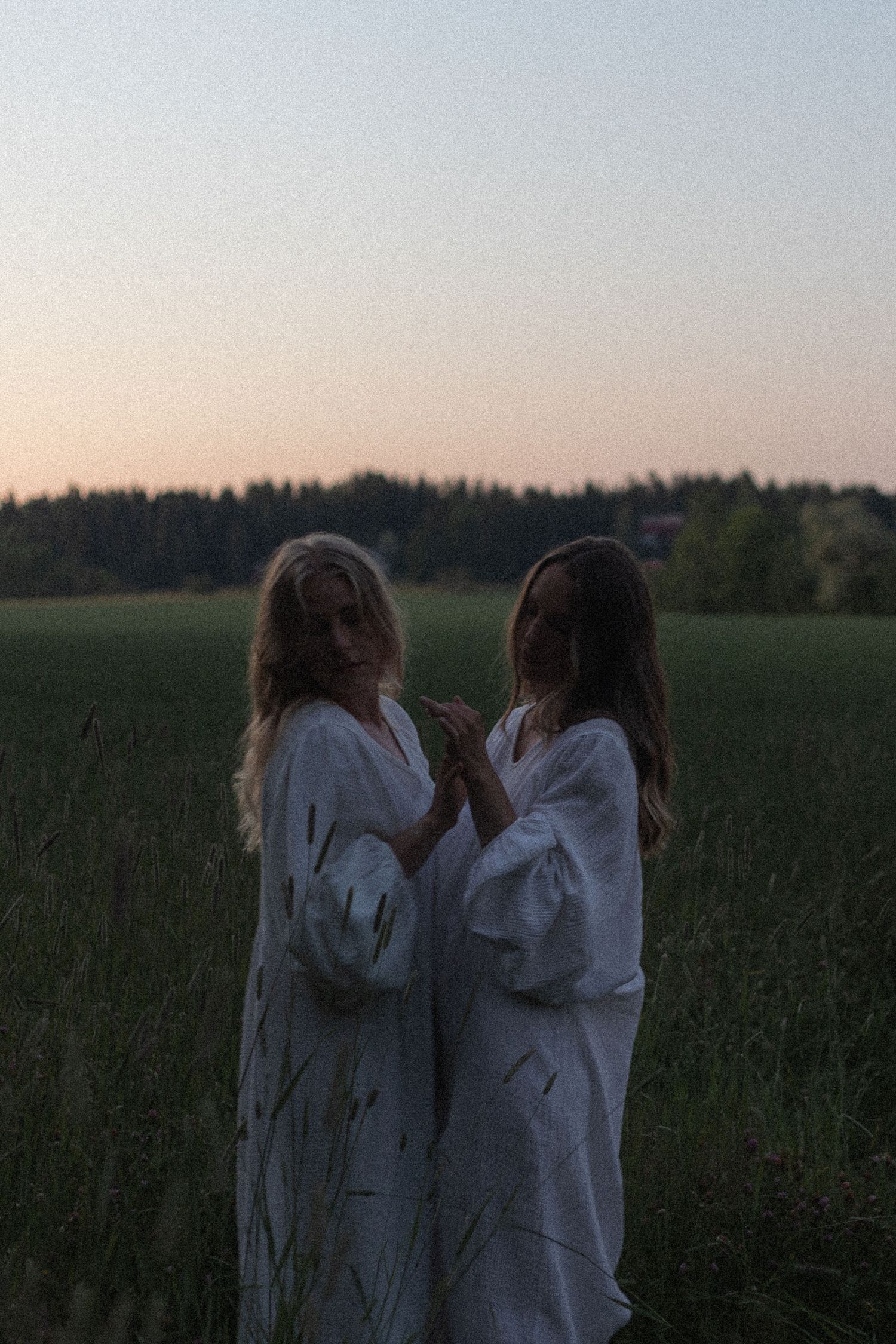 Kaksi valkoisiin mekkoihin pukeutunutta ihmistä seisoo yhdessä niityllä iltahämärässä, ja horisontissa näkyy puiden siluetti.