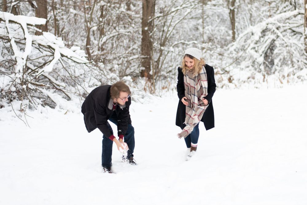 A playful winter scene of a couple bending down to make snowballs in a snow-covered forest.