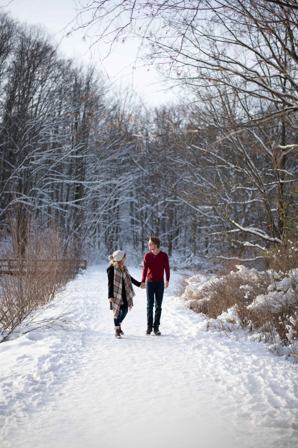 A couple holds hands while walking down a snowy trail surrounded by frost-covered trees.