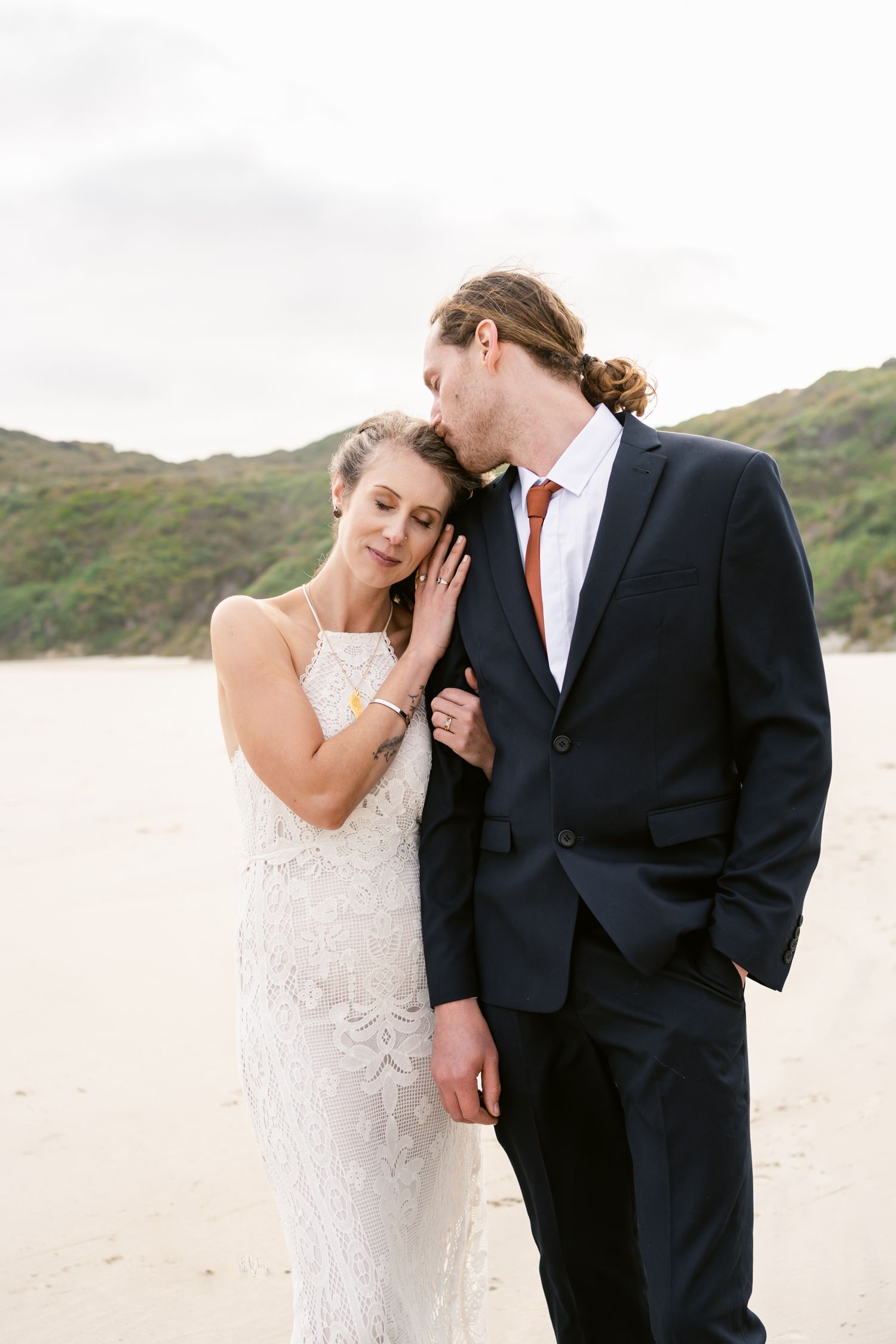 A bride and groom share an intimate moment on a beach with green hills in the background.