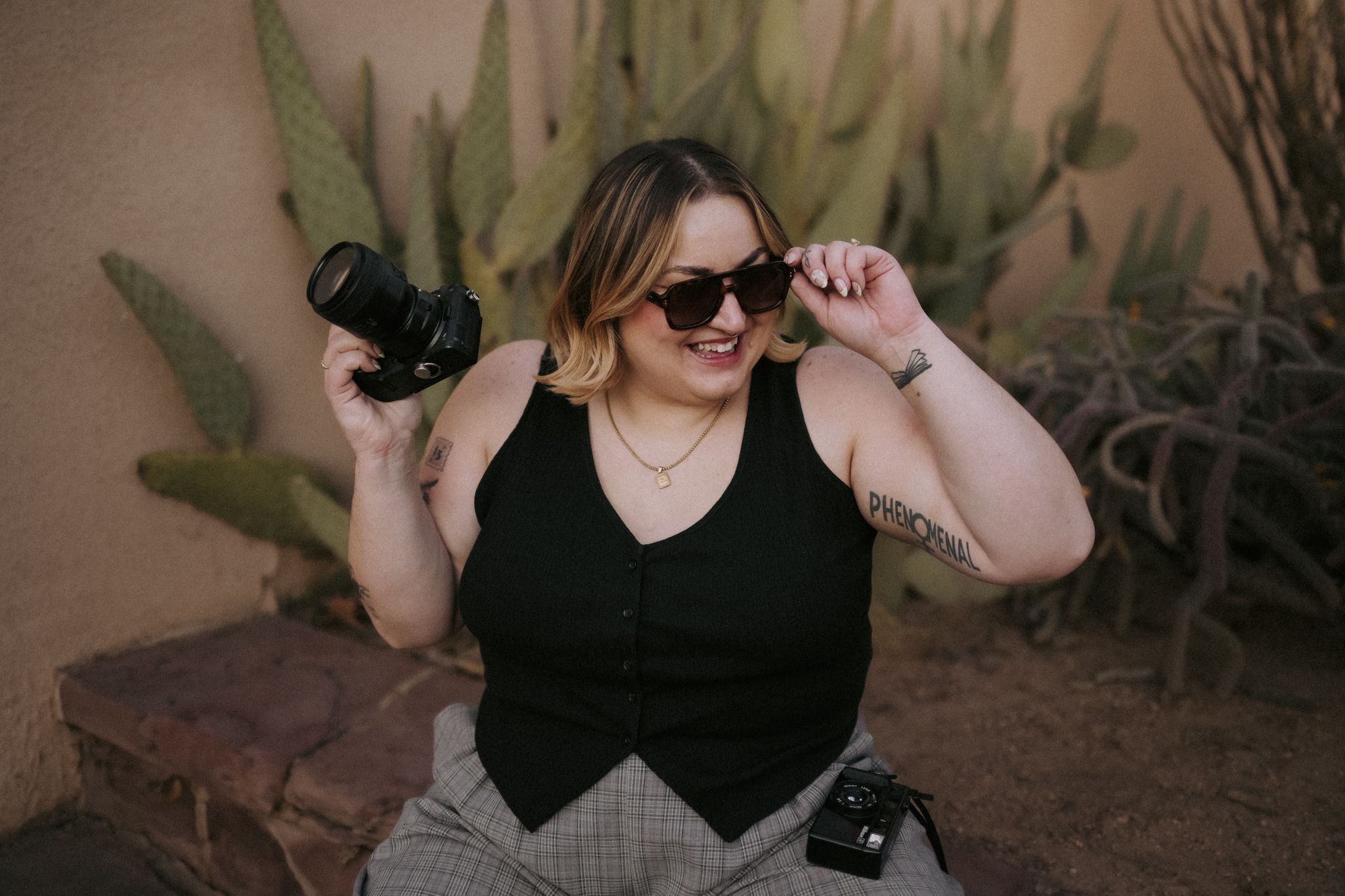 Professional photographer in black vest and sunglasses poses with camera equipment against desert plants backdrop. ERIKA DURDLE PHOTOGRAPHY