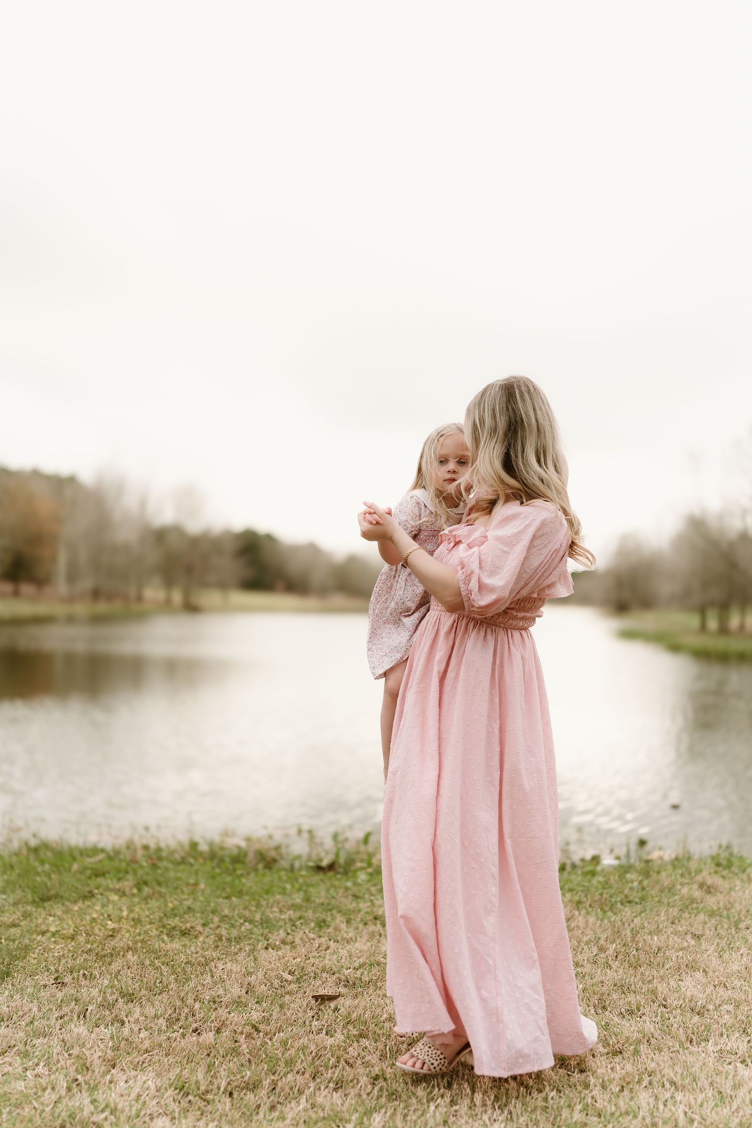 Two figures in flowing pink dresses embrace by a serene lake.