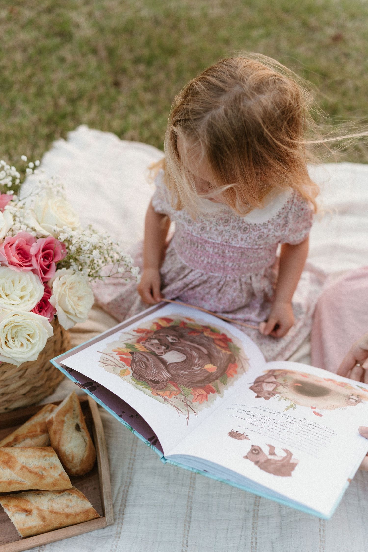 Reading a storybook together on a white blanket with pink roses and pastries nearby.