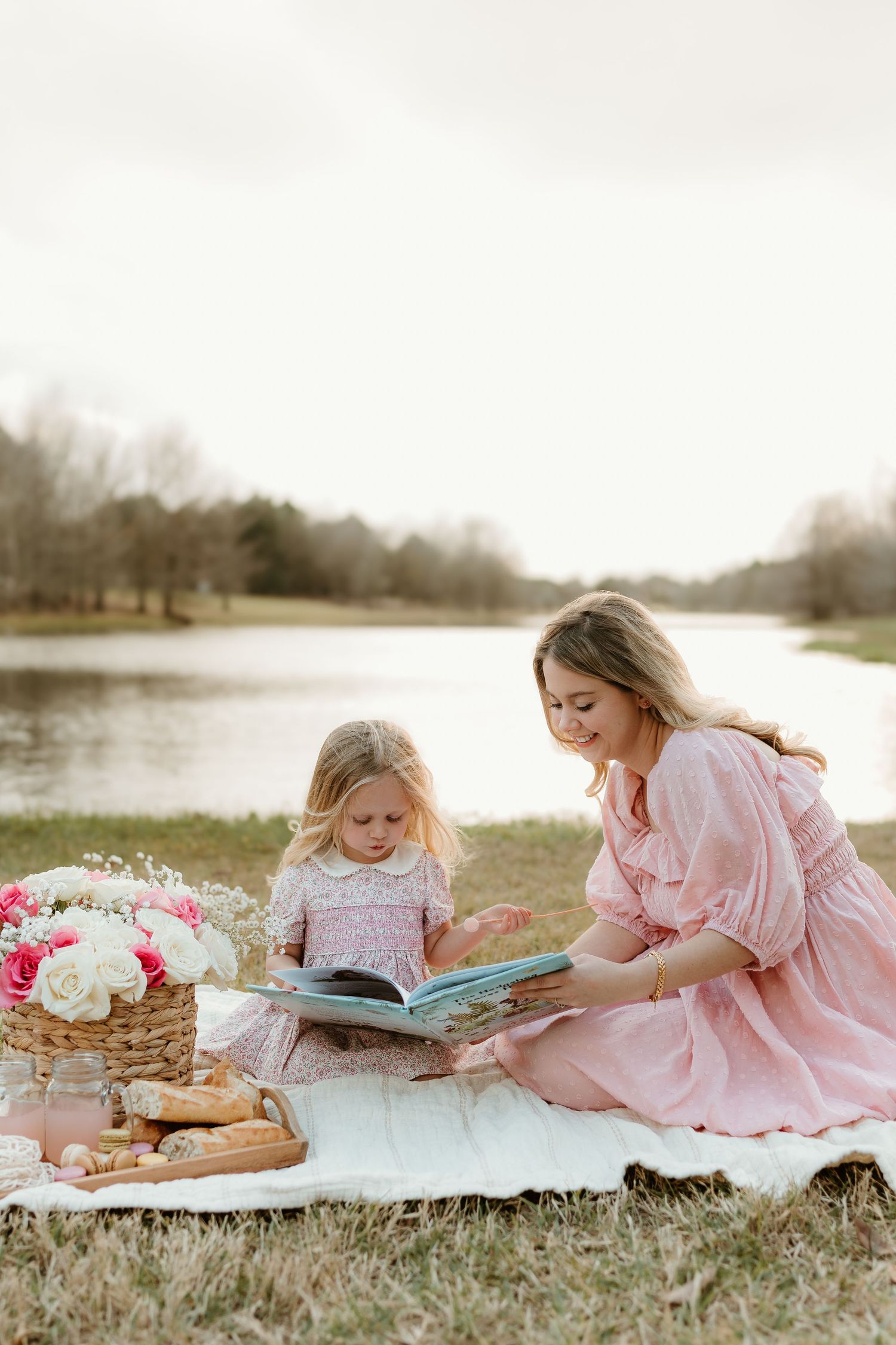 Cozy picnic scene with books, flowers, and two people in pink dresses reading by the lake.