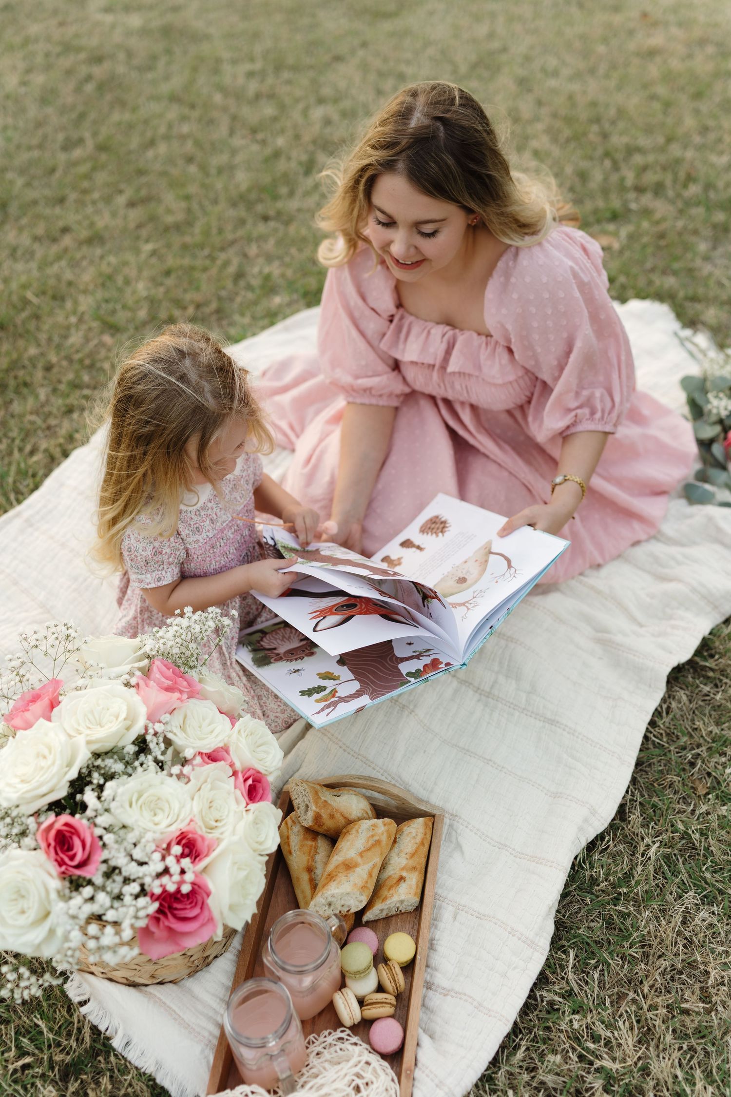 A picnic scene with pink dresses, flowers, and a book laid out on a white blanket in the grass.