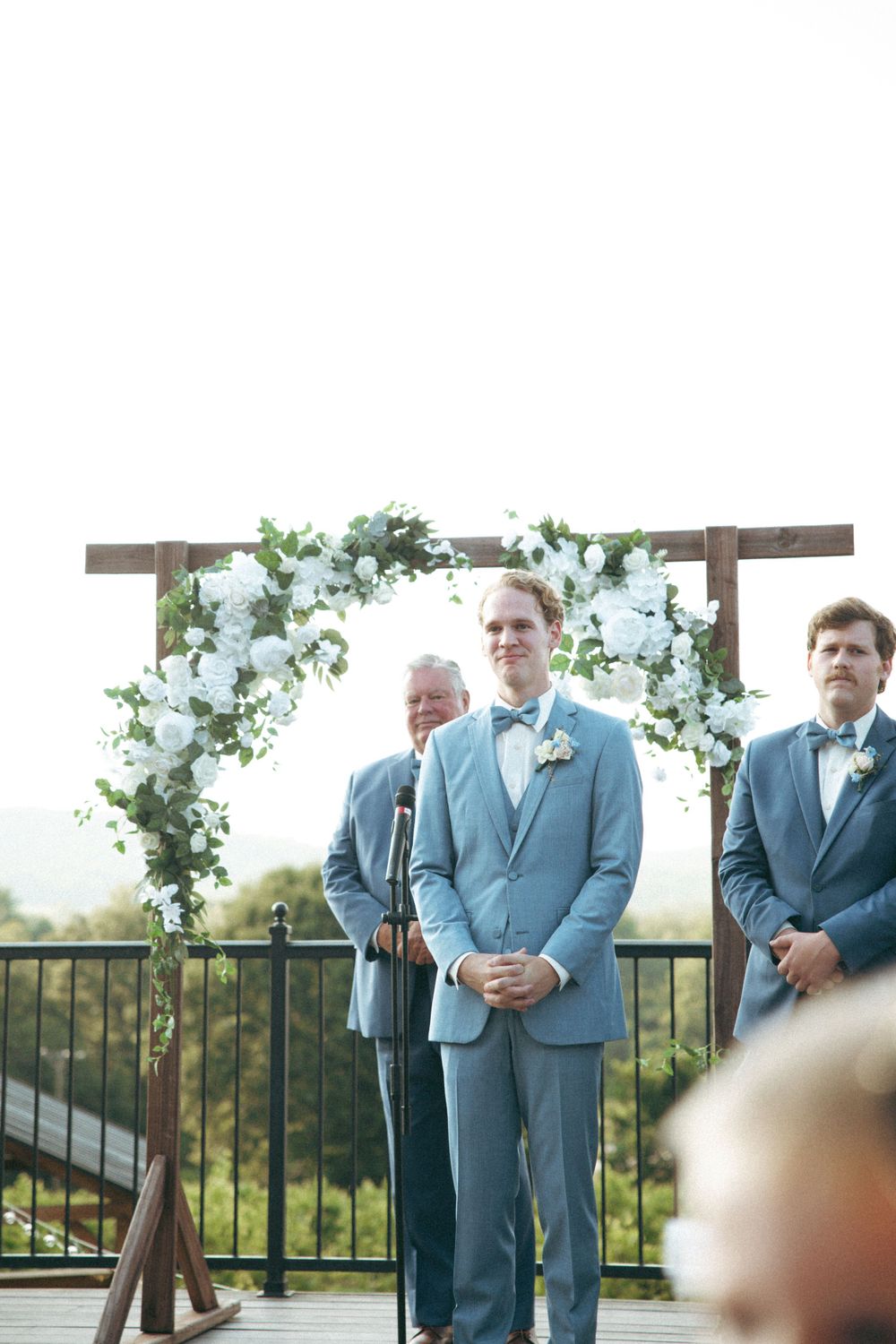 Wedding party stands at elegant outdoor ceremony arch decorated with white florals and greenery.
