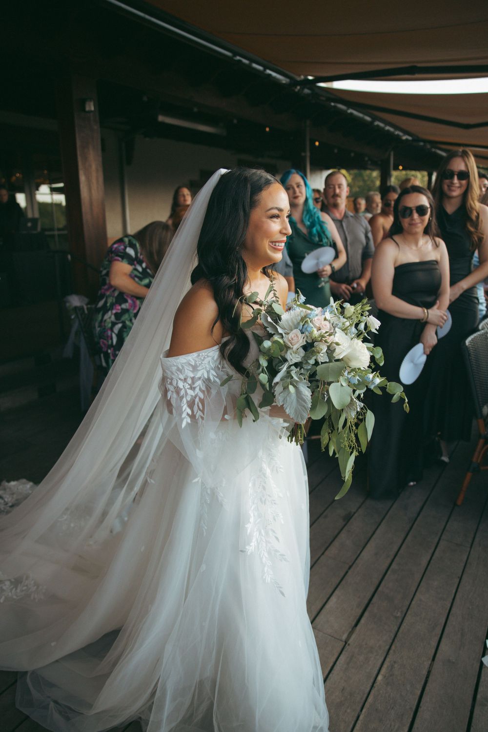 Bride in lace wedding gown carries white bouquet while smiling radiantly on wooden deck.